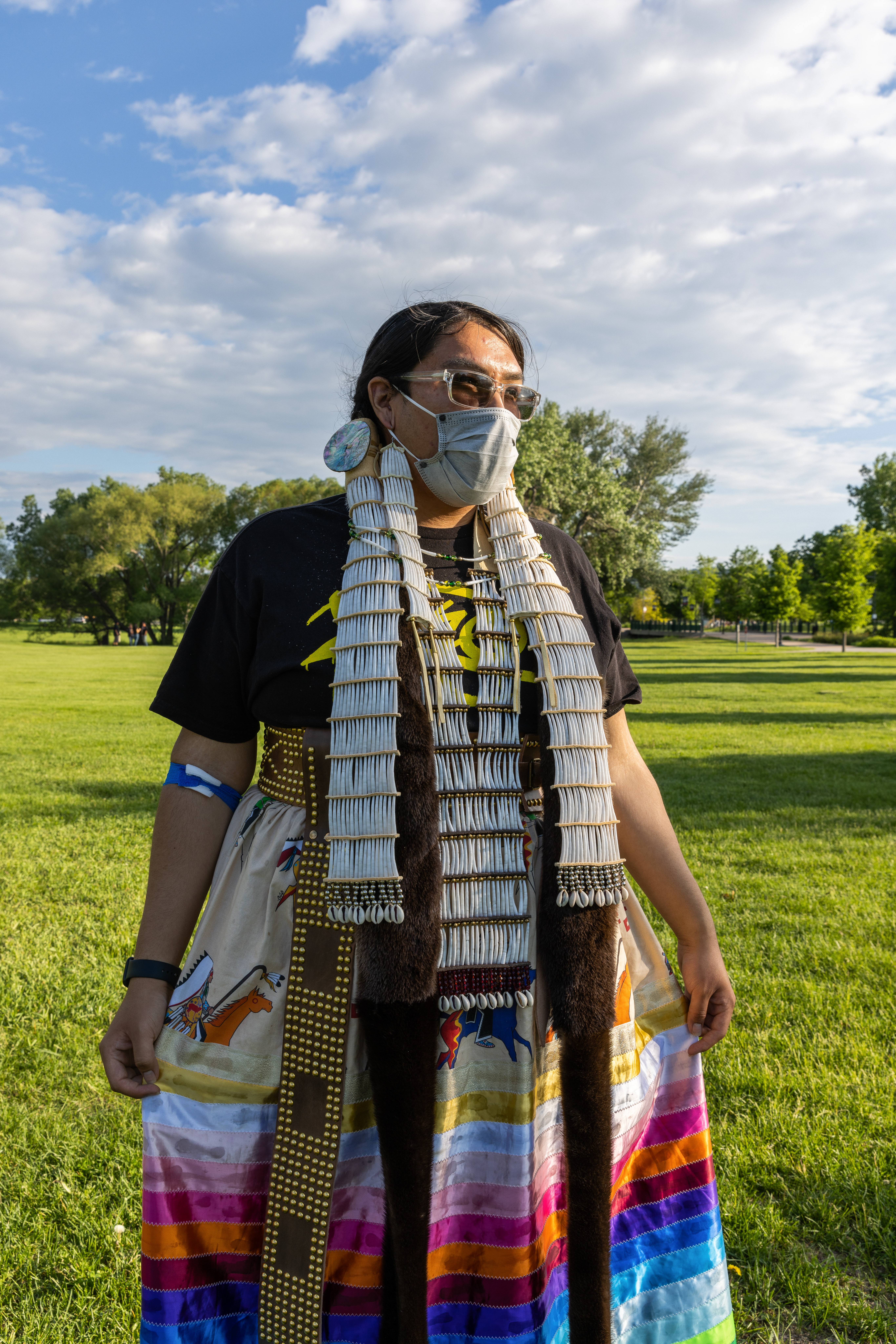 A person with medium-light skin tone wearing sunglasses, a cloth mask, and a beaded hairpiece and earrings, with a brown skirt and colorful skirt.