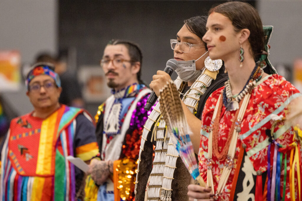 In a group of people all wearing colorful regalia, one person holding a microphone and speaking.