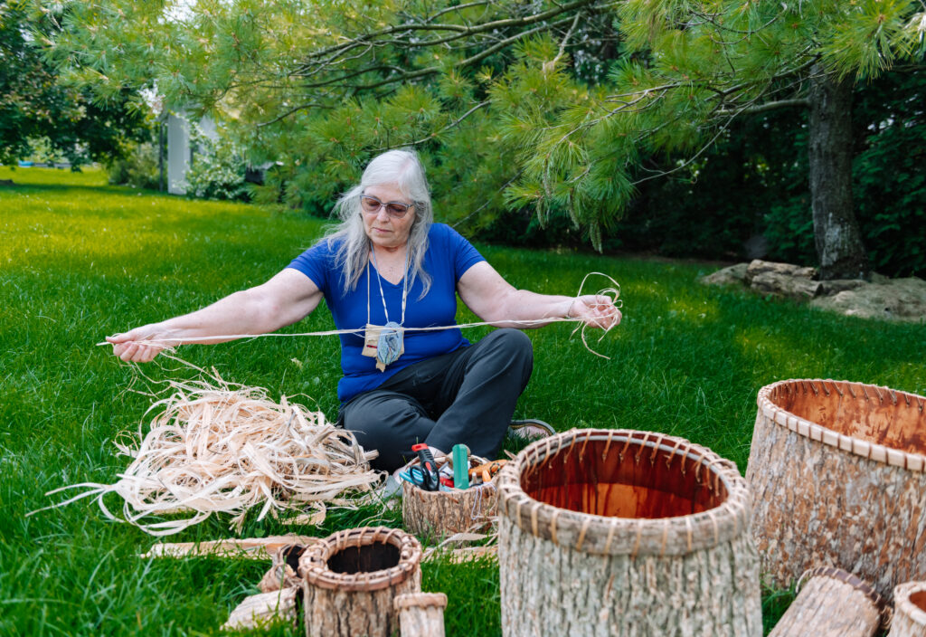 A person of light skin tone and long grey hair sits on green grass as they work with materials to make a wooden basket. They have their legs crossed and their arms stretched out as they hold what looks like a long, thin rope-like material.