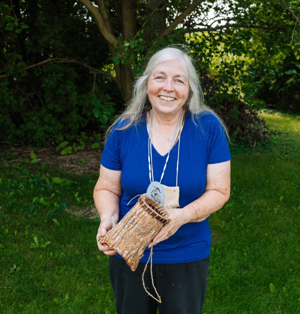 A person of light skin tone and long grey hair smiles and holds a small handmade basket made of tree bark. They are wearing a bright blue T-shirt and black pants.