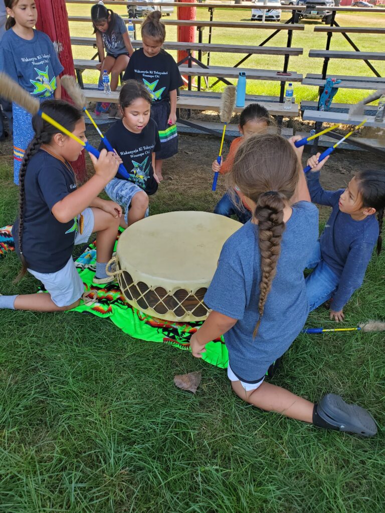 A group of half a dozen young children kneel around a drum, as they play the instrument and sing.