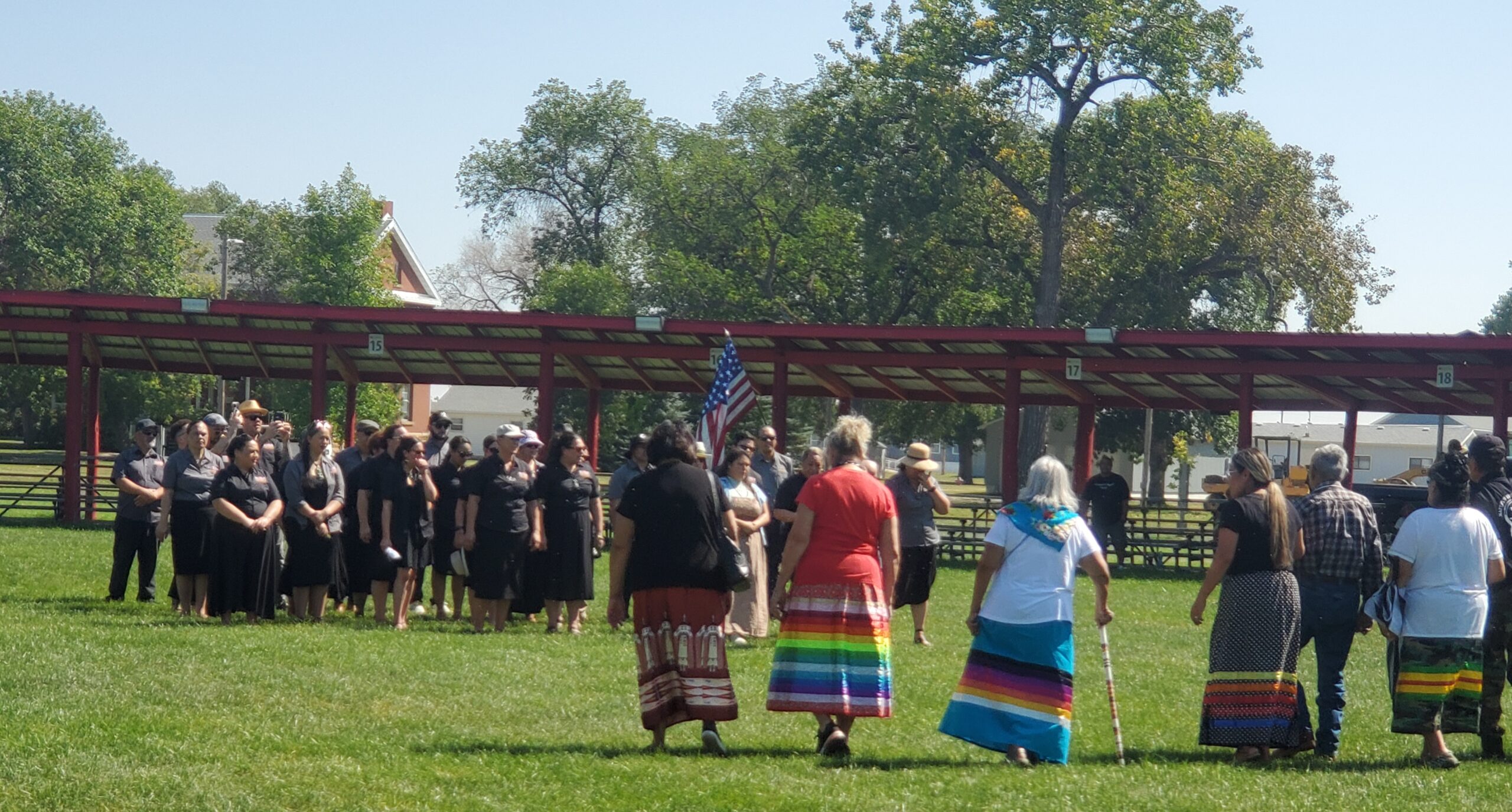 A group of people walk towards another group standing at a distance in an outdoor area. Some of them wearing colorful ribbons skirts, while others are dressed in black.