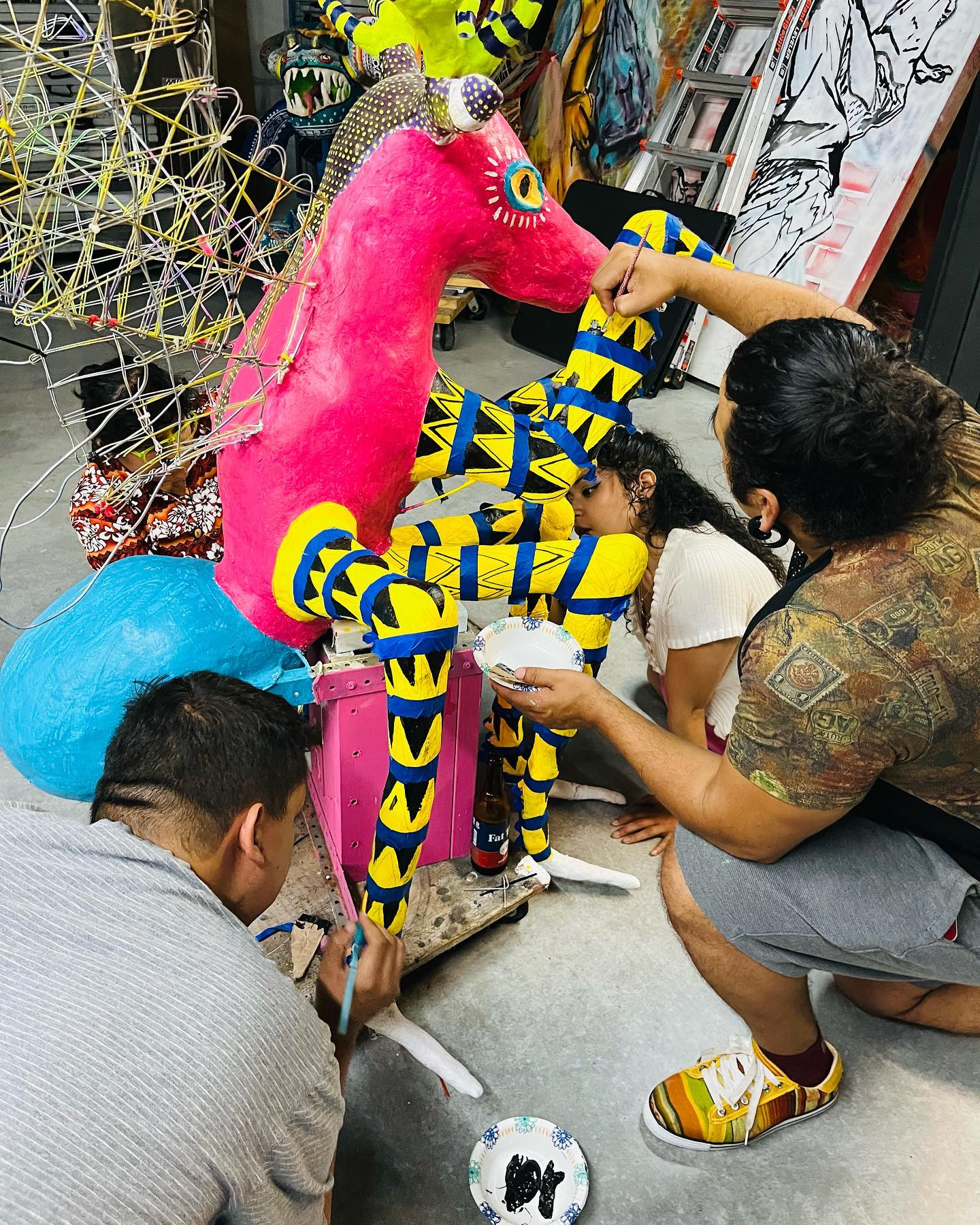 People with medium skin tone and dark hair collaborate on painting a colorful sculpture of an alebrije