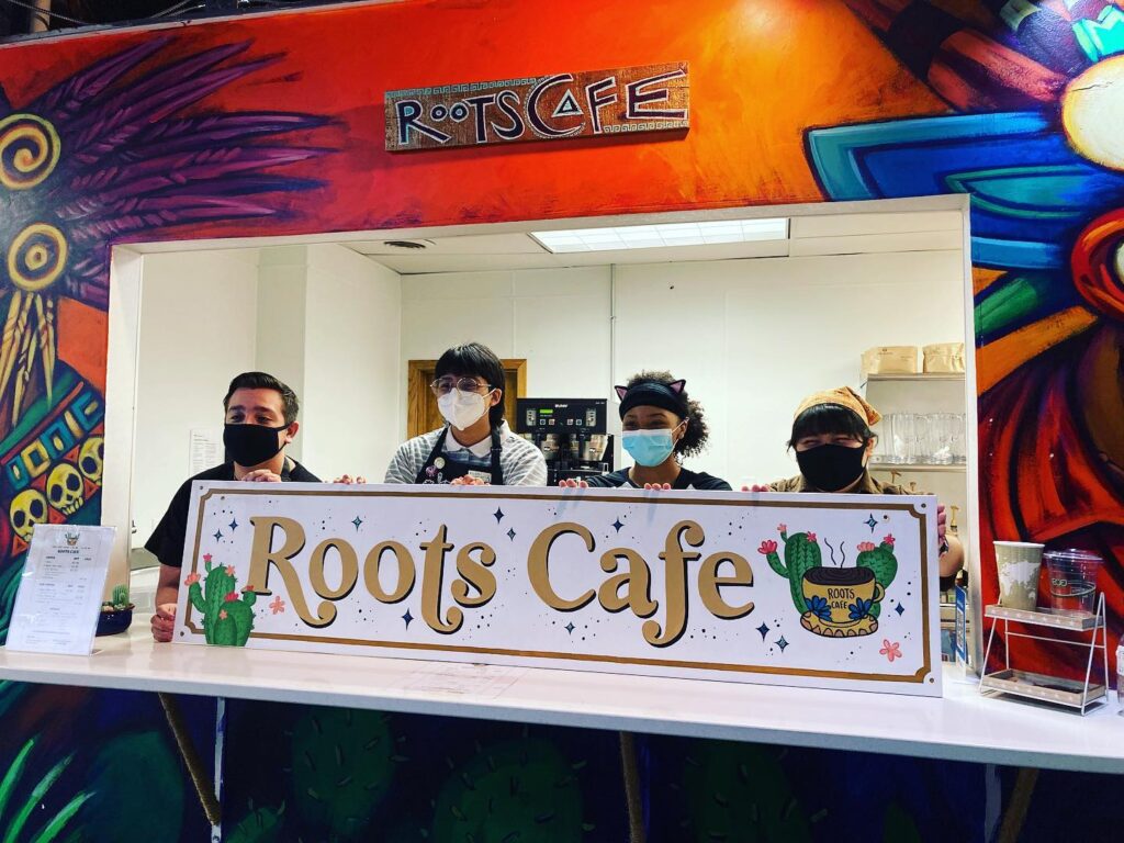 People stand in a kitchen behind a sign that reads "Roots Cafe"
