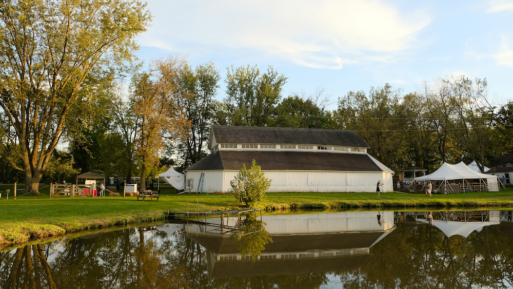 A view of a pond with a long barn and large tent beside it. There are trees and greenery around it.