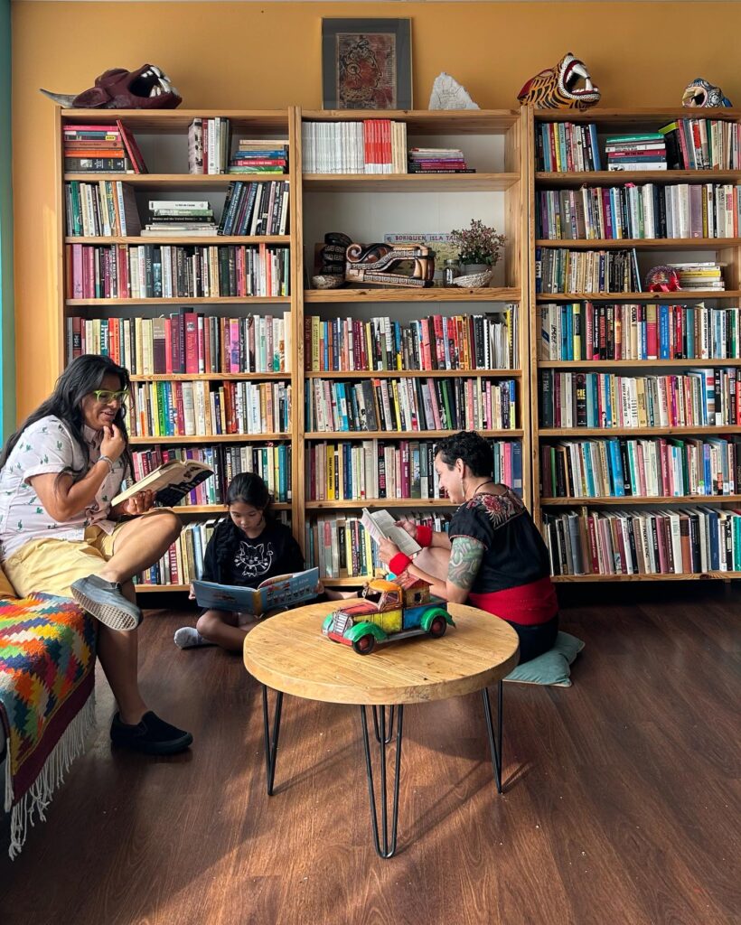 People sit in a group reading books. Behind them is a set of bookshelves filled with books, sculptures, and plants.