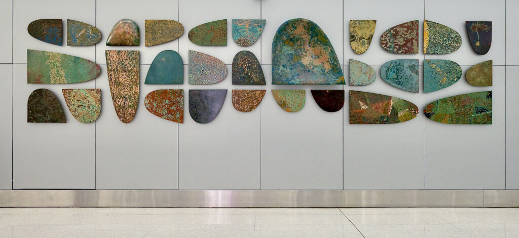 A wall with an assortment of three-dimensional works of varying sizes. They have abstract patterns made of a range of colors from yellows, greens, blues, and browns.