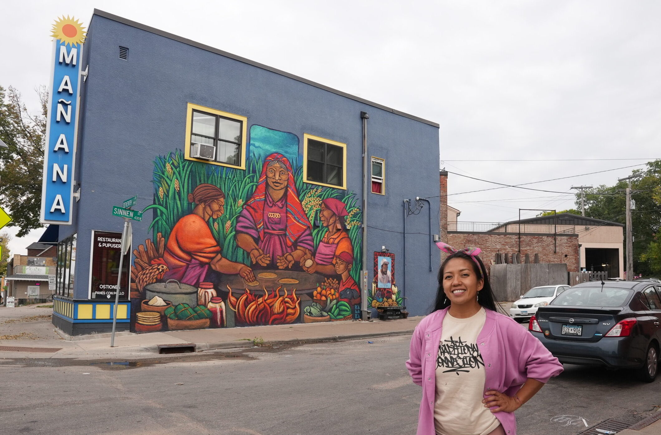 A person with a medium skin tone wearing a pink jacket stands in front of a mural of three people sitting around a hot surface rolling dough and cooking it. The mural is painted on a stucco wall of the Manana Restaurant & Pupuseria.