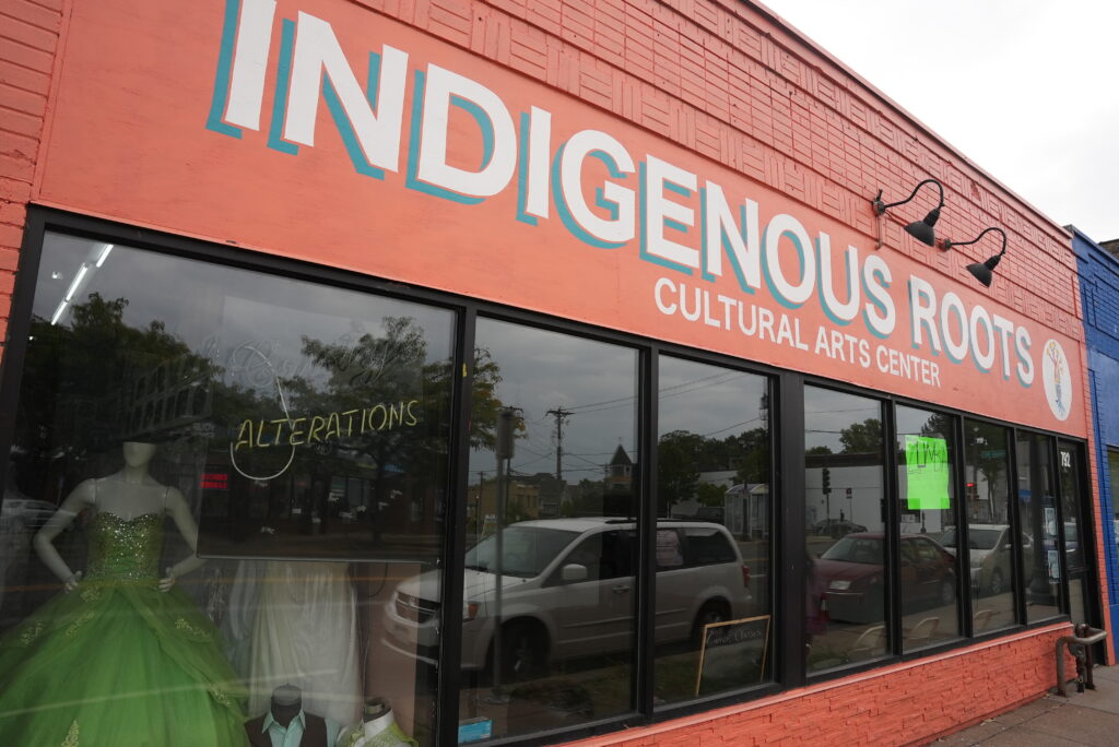 A bright orange building facade with text that reads Indigenous Roots Cultural Arts Center. In the window is a sign for alterations next to a bright green formal dress.