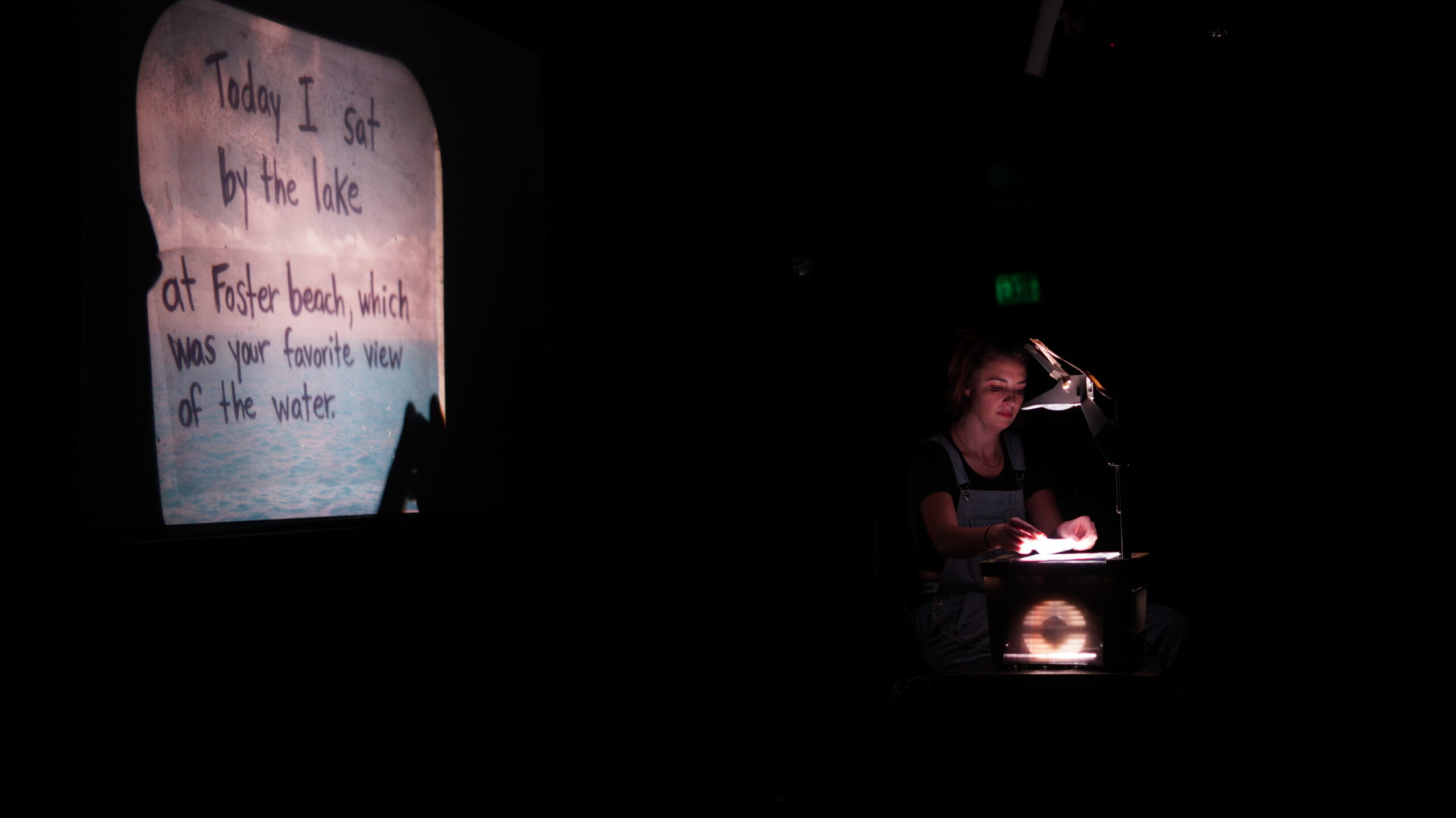 A person of light skin tone sits by an overhead projector, while a screen behind them shows an image of a beach with words that read, "Today I sat by the lake at Foster beach, which was your favorite view of the water."