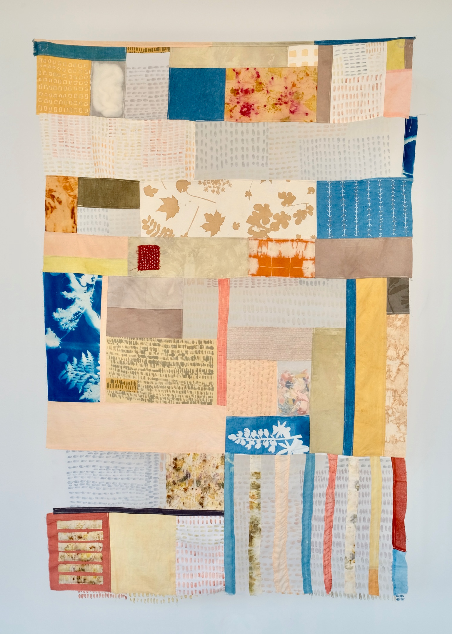 A patchwork quilt hung on a white wall. The quilt is made of naturally dyed fabrics in shades of yellows, reds, blues, and browns.