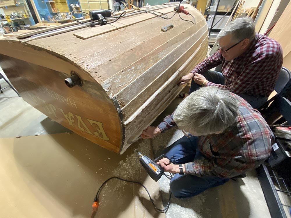 Two adults hunch over an old boat as they work on it with hand woodworking tools and an electrical drill.