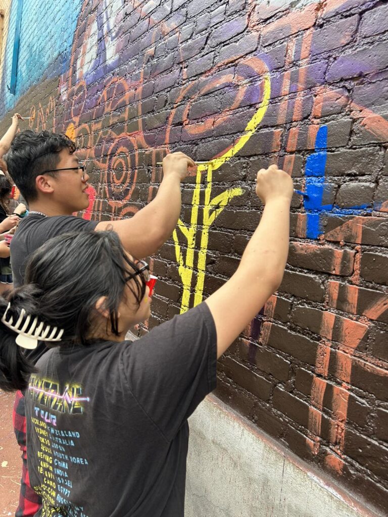 Two individuals painting a section of a mural on a brick wall