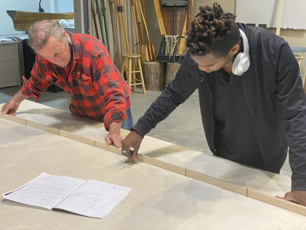Two adults measure and mark out a wooden piece during a boat building class.