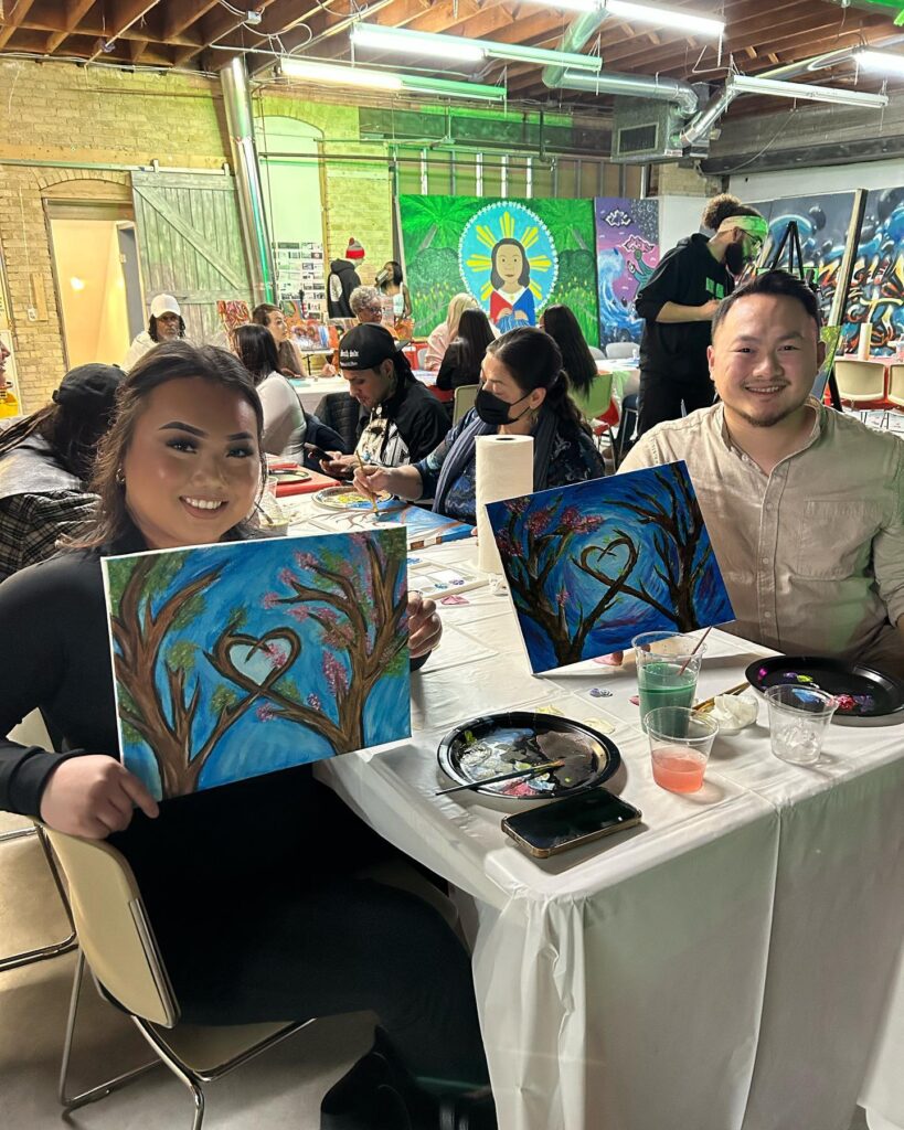 A room is filled with people painting a similar image. Two people show their finished work which includes two trees whose intertwined branches make a heart shape.