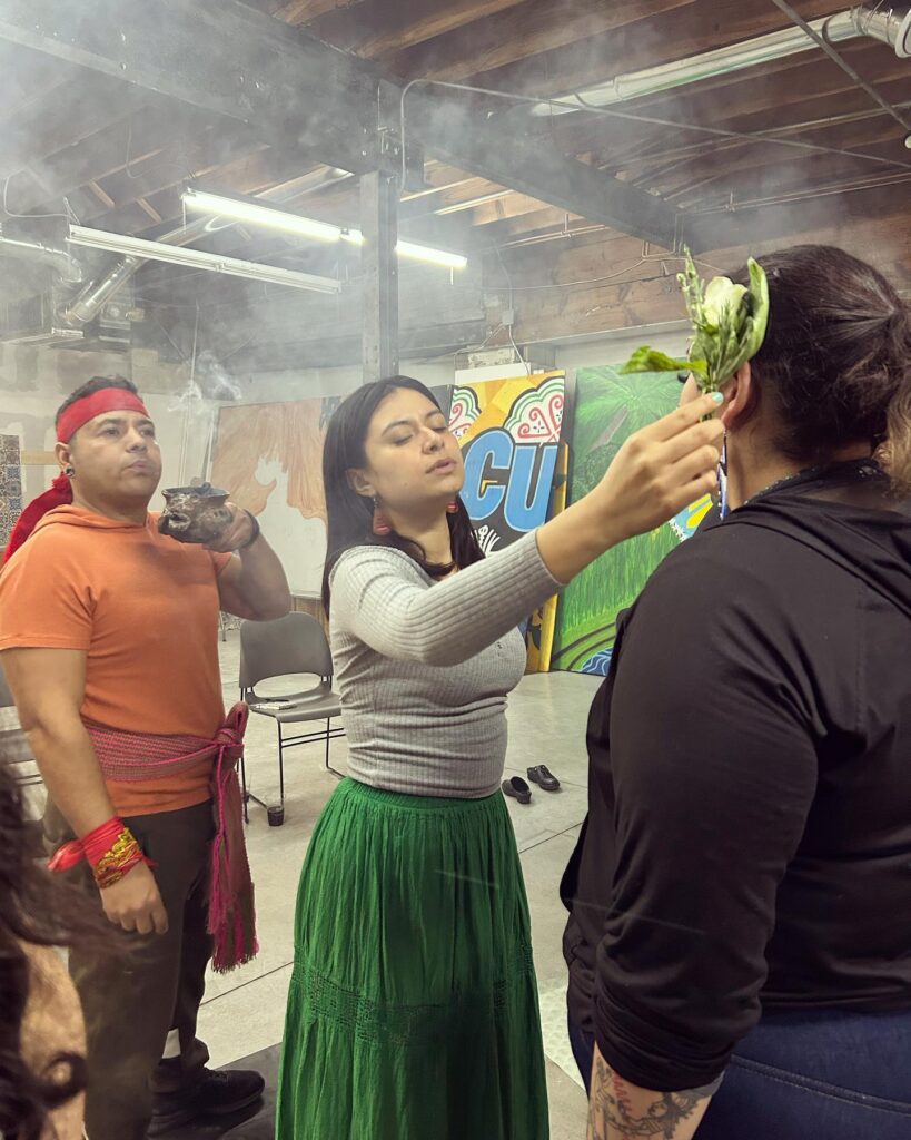 A person in a green skirt holds a plant to the side of another person's head. Behind them, someone holds a bowl emanating smoke and gently blows it forwards.