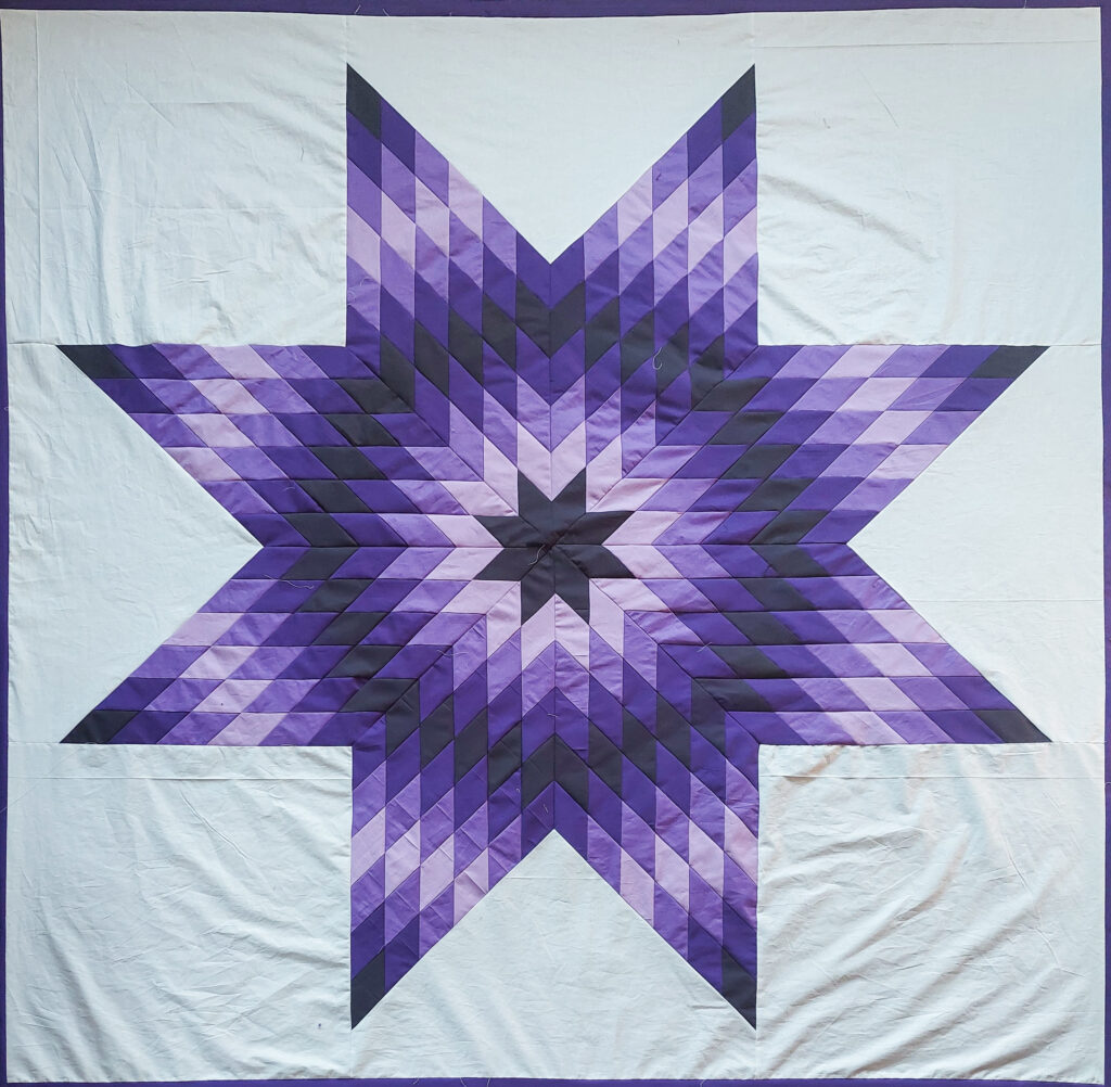 A quilt with an eight-point star pattern made of deep and light purples on a white fabric.
