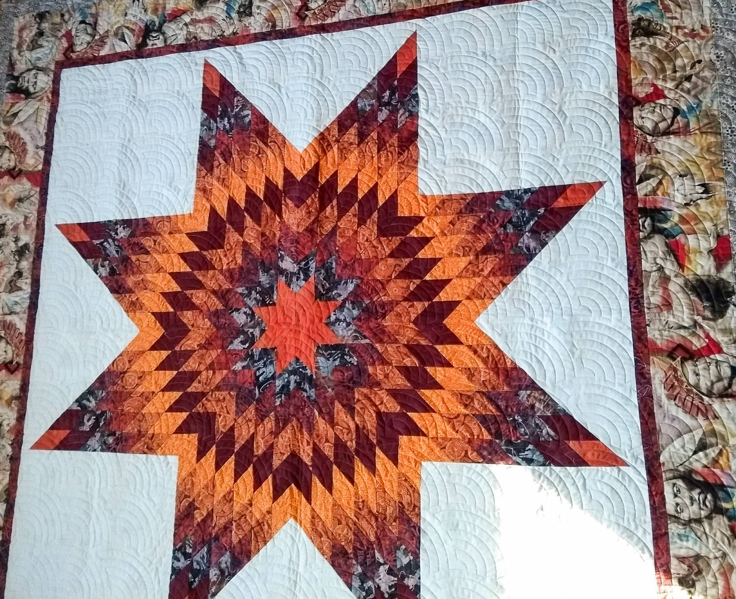 A quilt made with an eight-point star pattern with shades of orange and red and a white backdrop. The edge of the quilt is made of a patterned printed fabric depicting a person in Native American head gear.