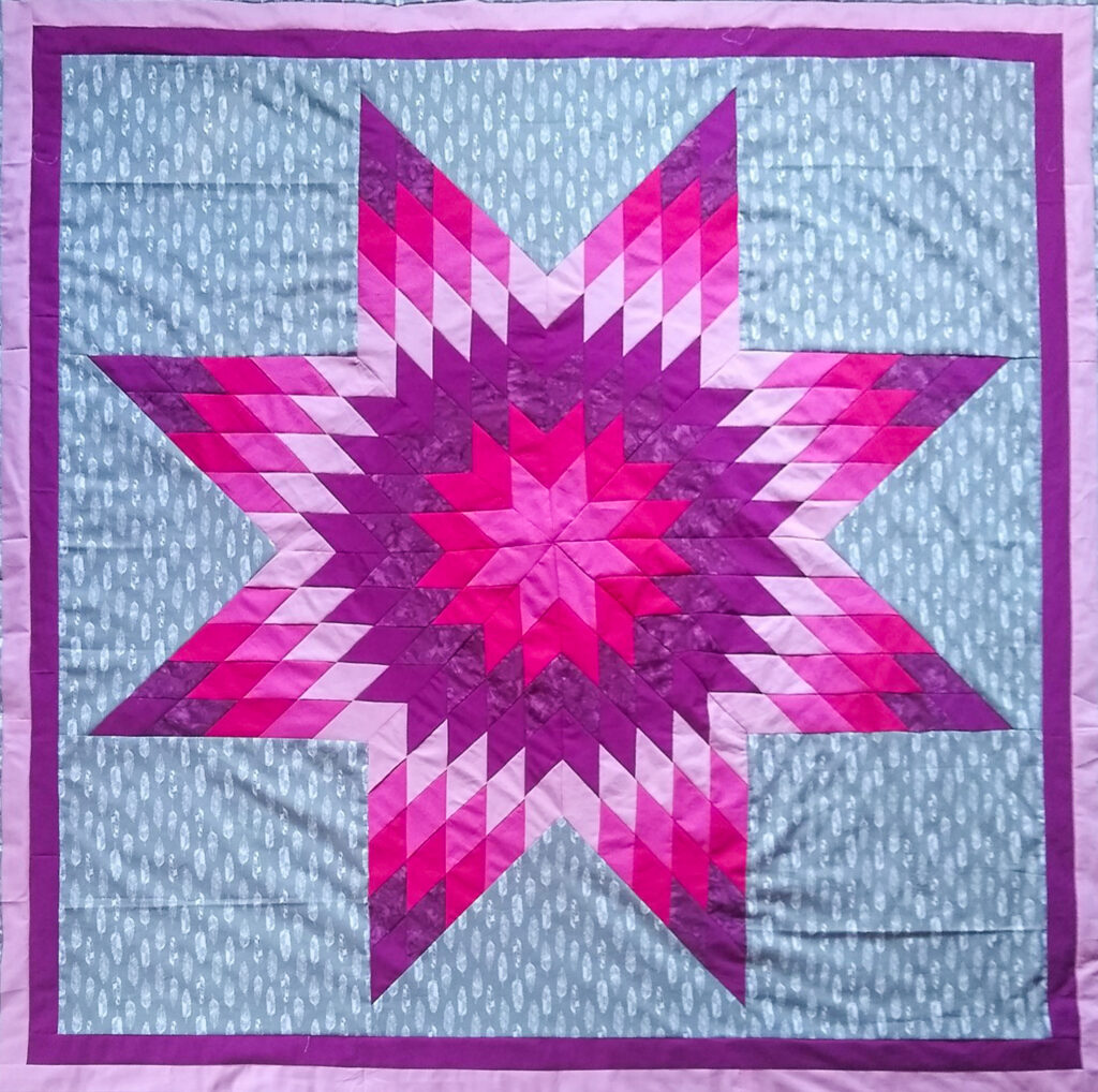 A quilt with an eight-point star pattern made of pinks, purples, and grey.