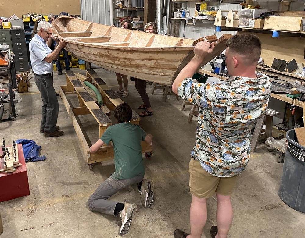 A group of adults hold up a wooden boat they are restoring, as one person adjusts a structure that the boat will be placed on. They are in a wood shop.