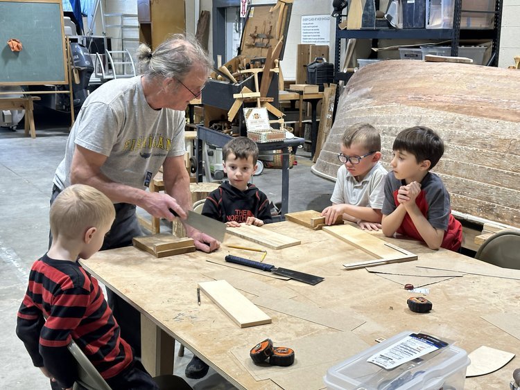 Four young children observe intently as their wood working instructor demonstrates a lesson with a hand-held wood saw and a wood block.