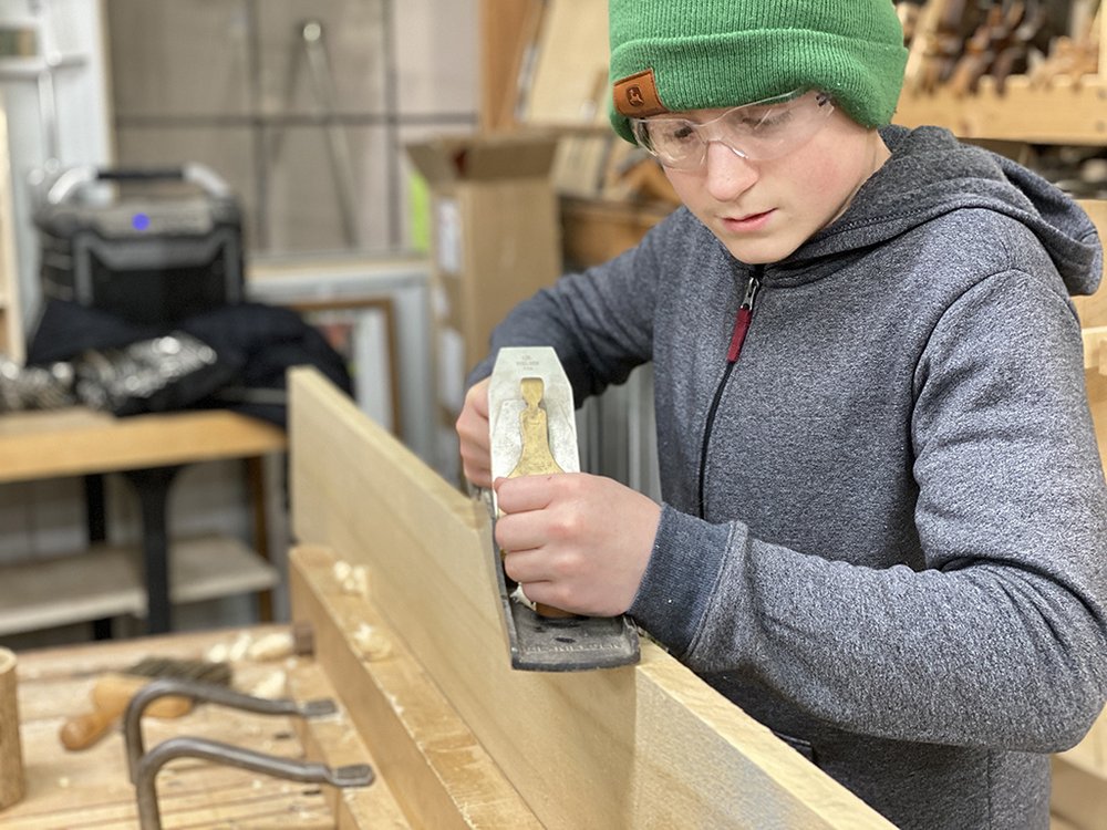 A young adult wearing a green beanie and safety glasses operates a handheld planer on a wooden piece.