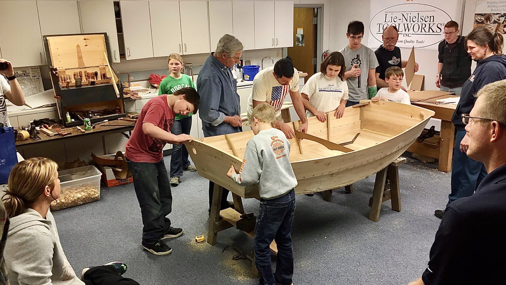 A group of children of varying ages huddle around an in-progress small boat during a boat building class. There's an adult supervising them, while others stand around and observe the process.
