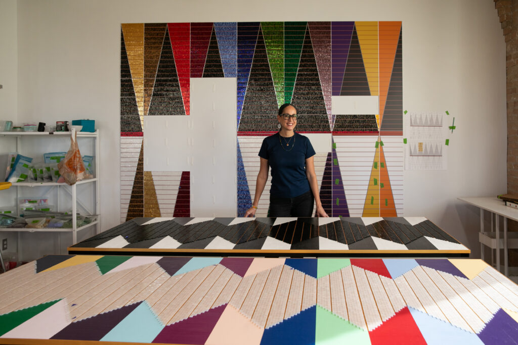 A person with dark hair and glasses stands at a table displaying colorful, geometric art. More artwork is shown in the background.