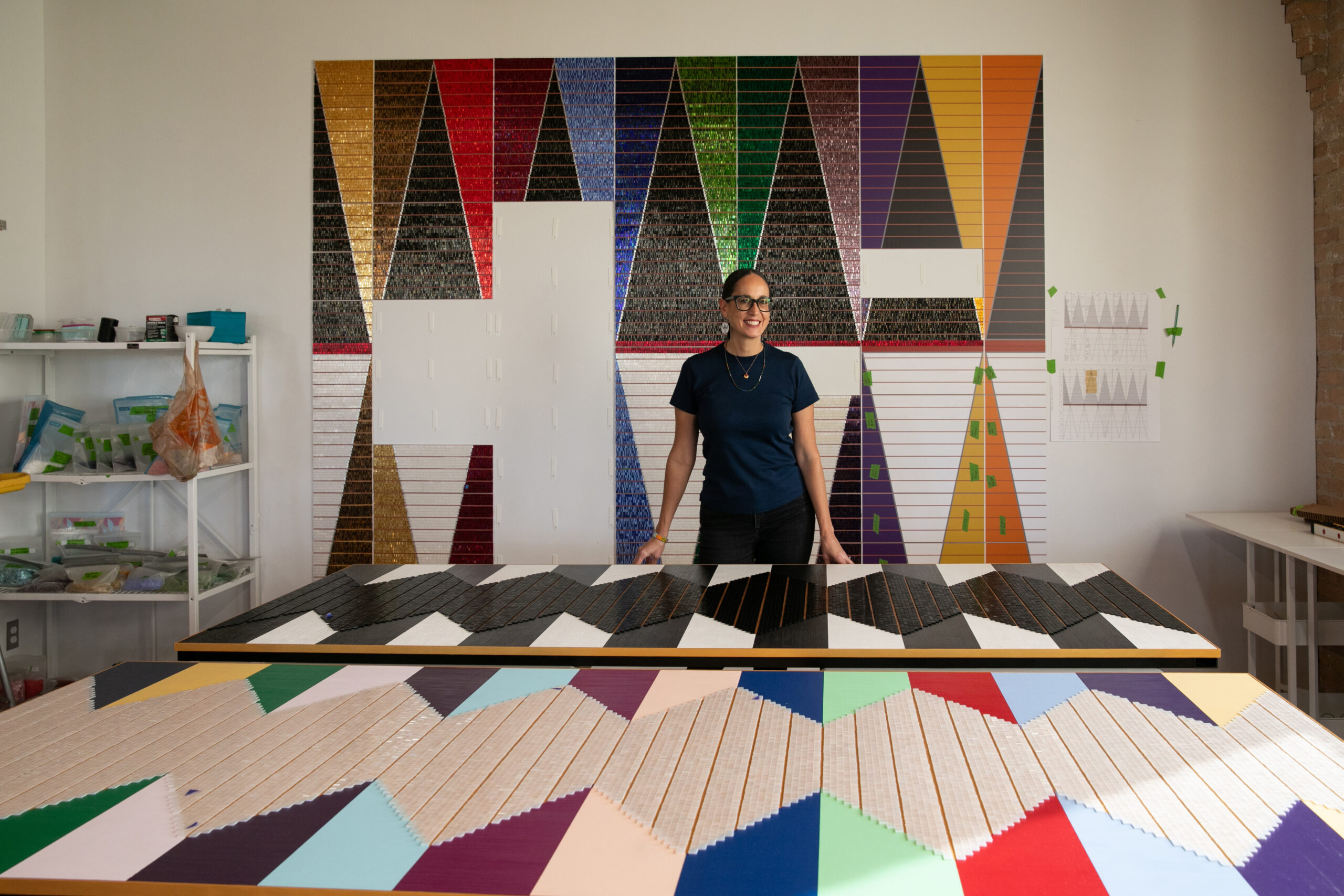 A person with dark hair and glasses stands at a table displaying colorful, geometric art. More artwork is shown in the background.