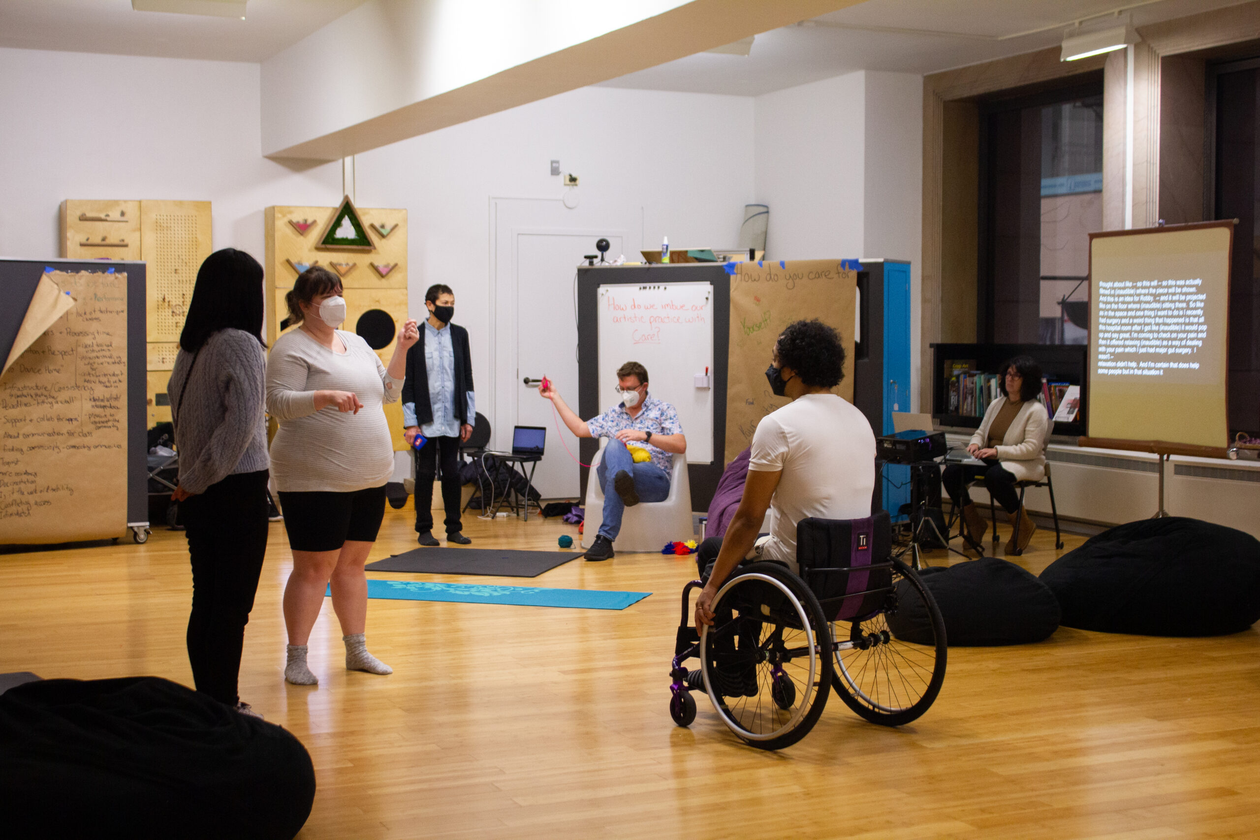 A group of disabled dancers in masks convene in an indoor space with wood floors.