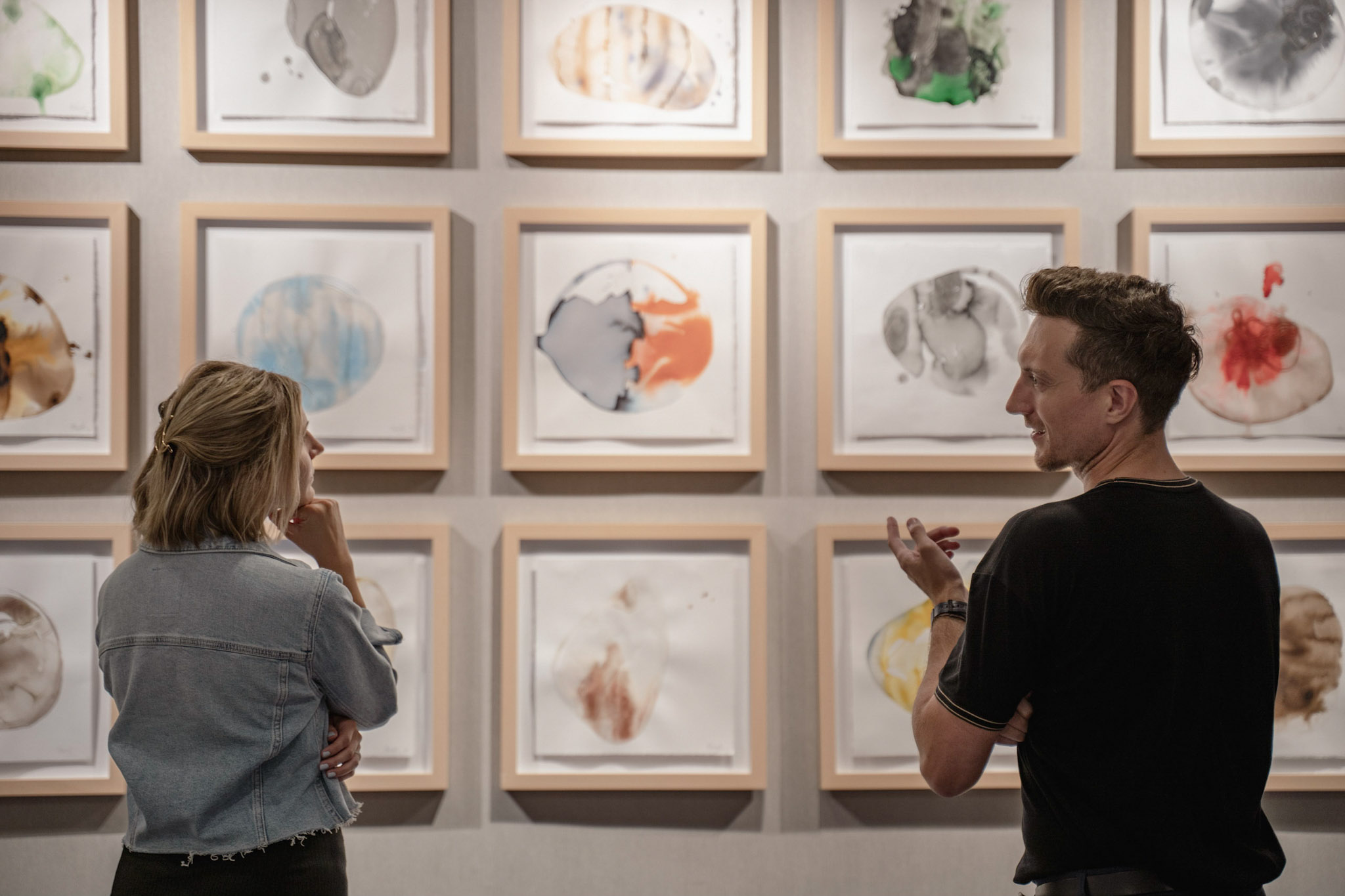 Two people standing in front of a wall of framed paintings deep in conversation.