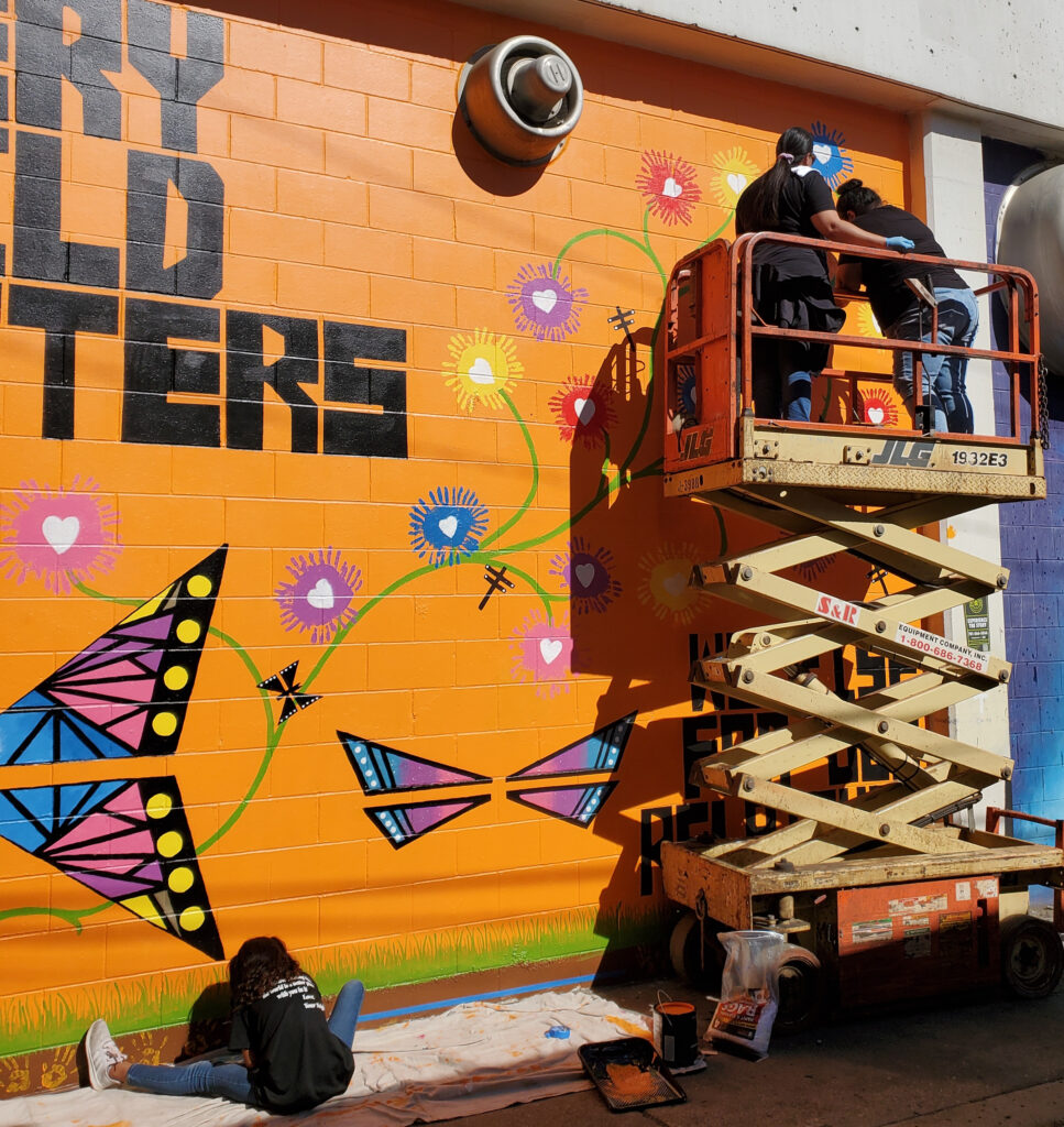 Two people wearing black T-shirts and blue jeans on a scissor lift, working a mural with a bright orange backdrop and depictions of flowers, butterflies, and dragonflies.