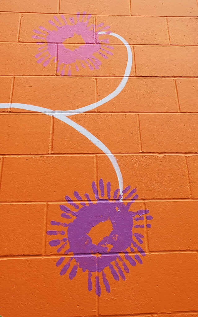Multiple handprints in purple paint made to depict a flower-like image on a bright orange wall.