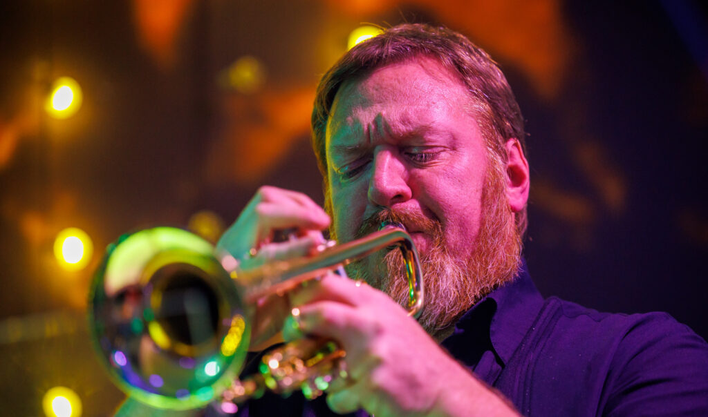 A person with light skin plays a trumpet under magenta stage light.