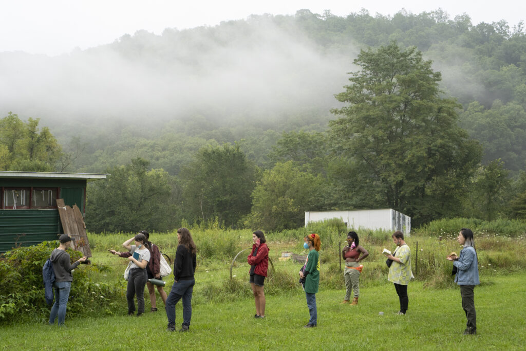 A group of people stand on green grass and listen to someone present near a large plant. Mist covers the treetops in the distance.