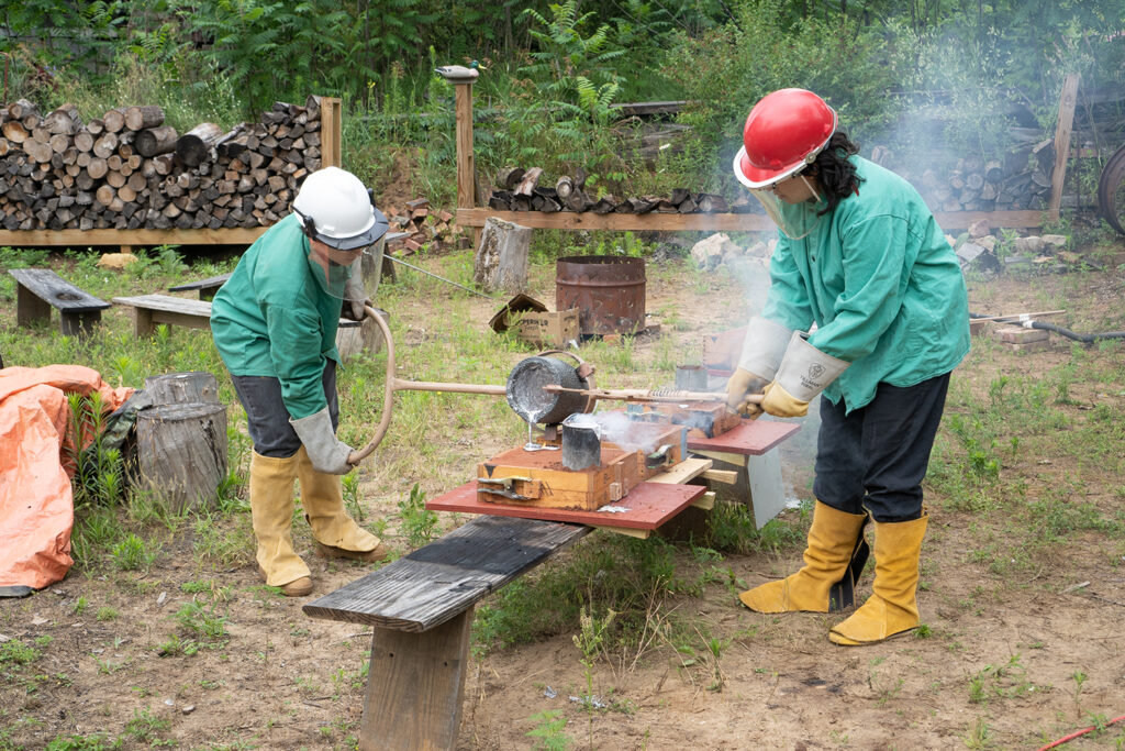Two people in protective gear pour hot melted metal into a mold.