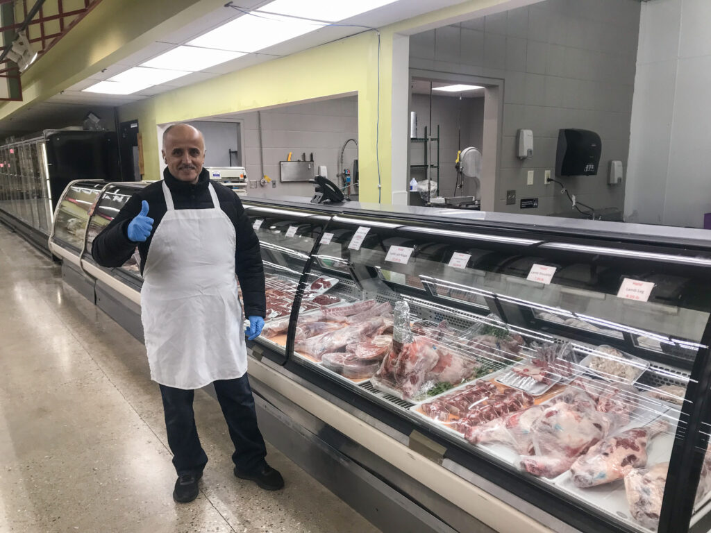 A man in an apron and gloves poses with thumbs up to the camera in a butcher shop