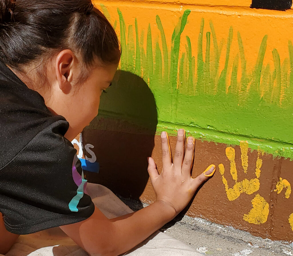 A child adds their handprint to a mural wall using yellow paint.
