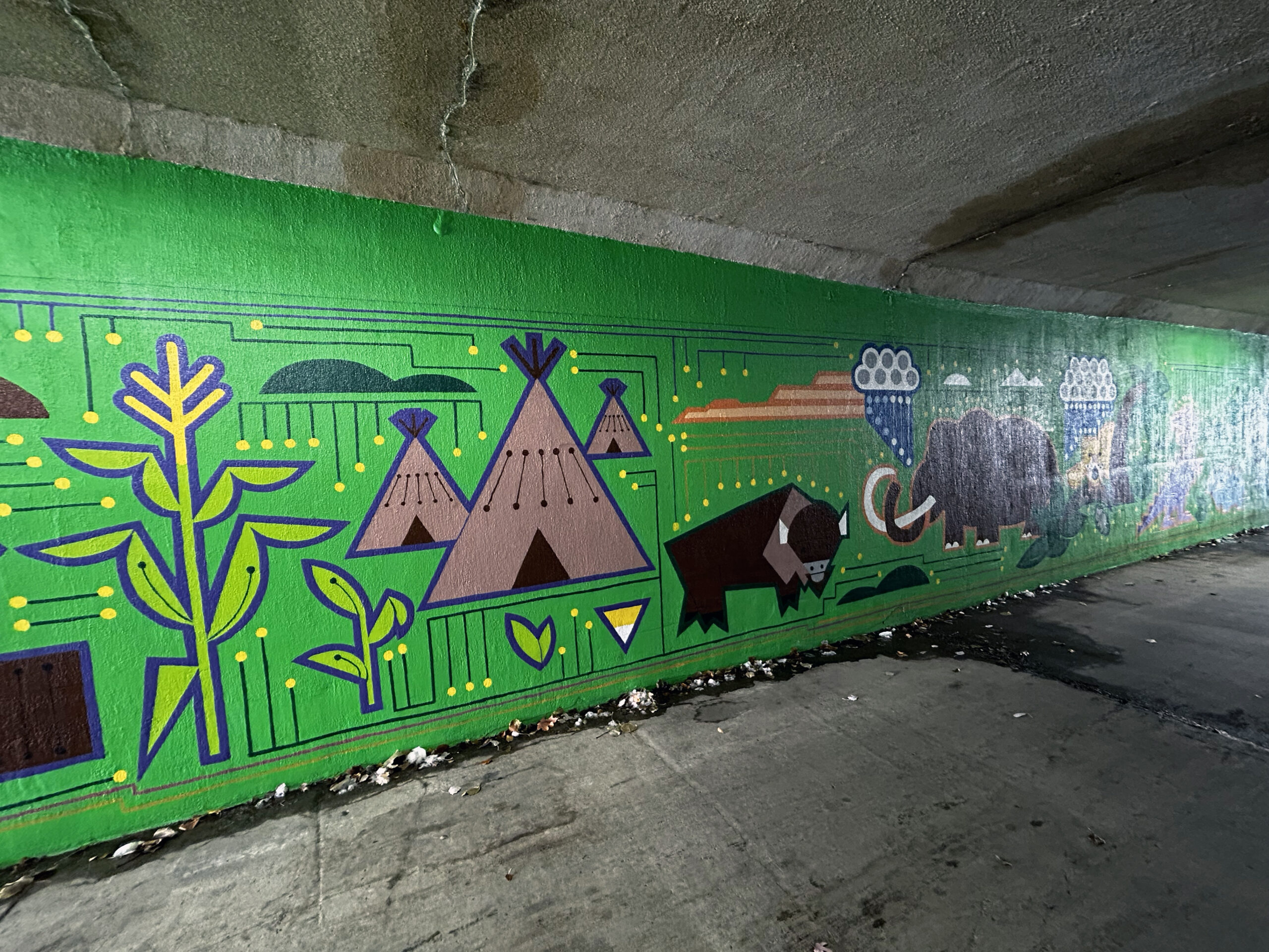A green mural under a bridge showing corn, teepees, and buffalo