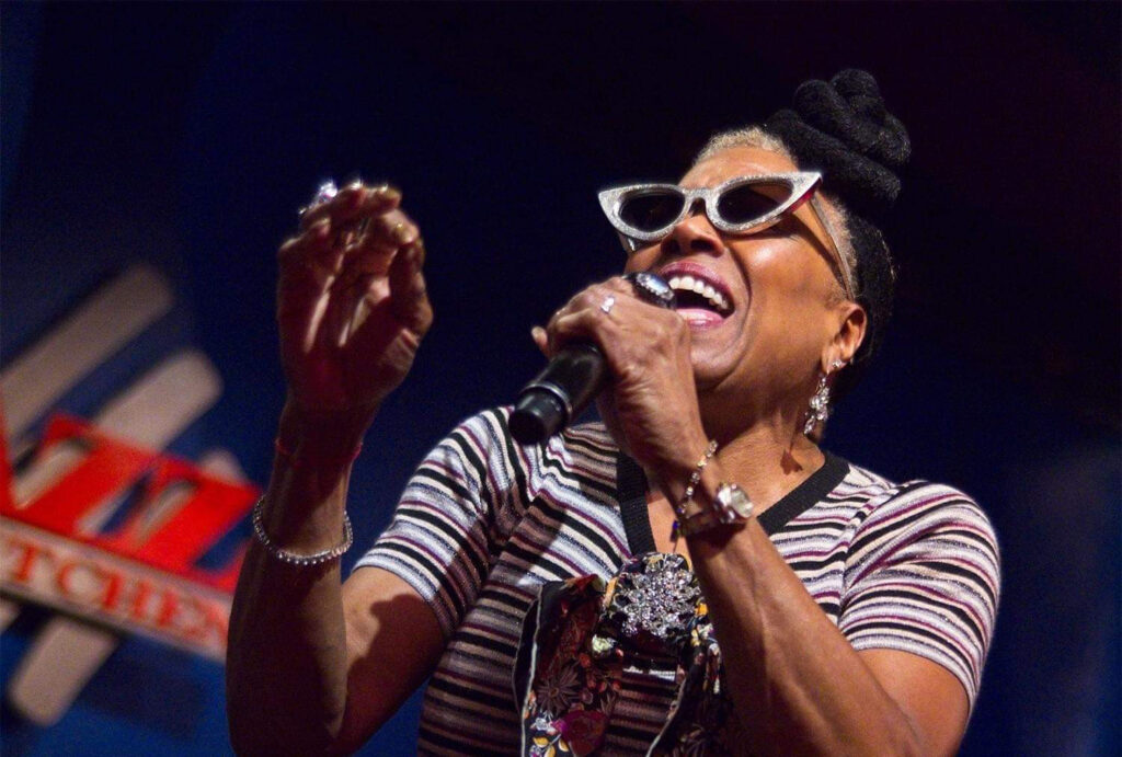A woman with medium-dark skin sings into a microphone. She wears silver metallic cat-eye sunglasses.