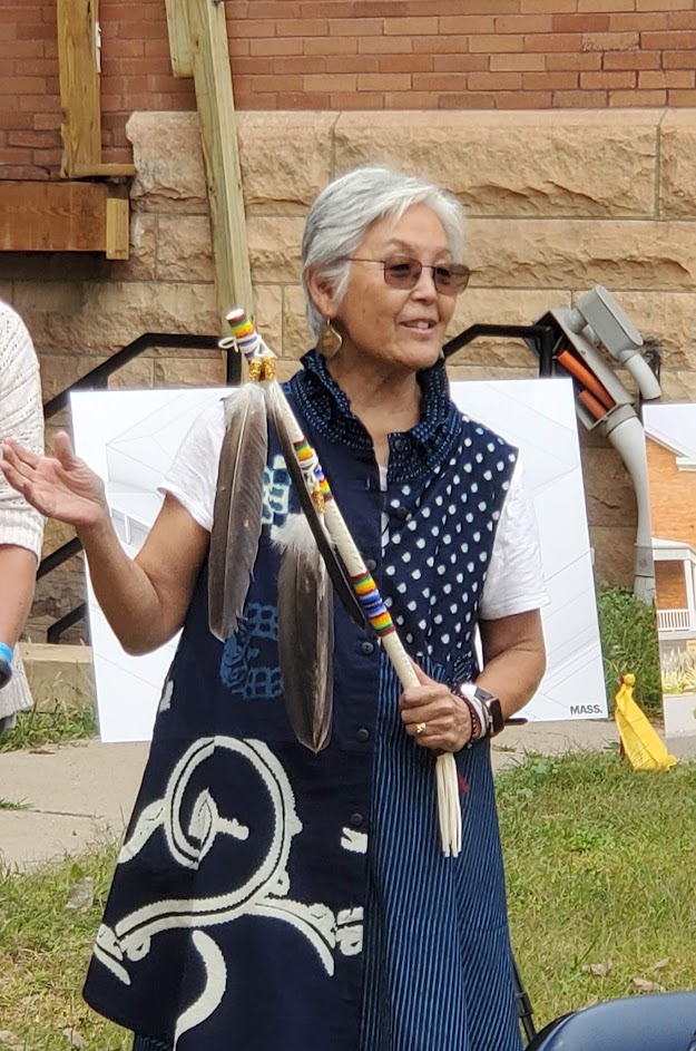 A Japanese American woman stands outside and speaks with a ceremonial object in her hand