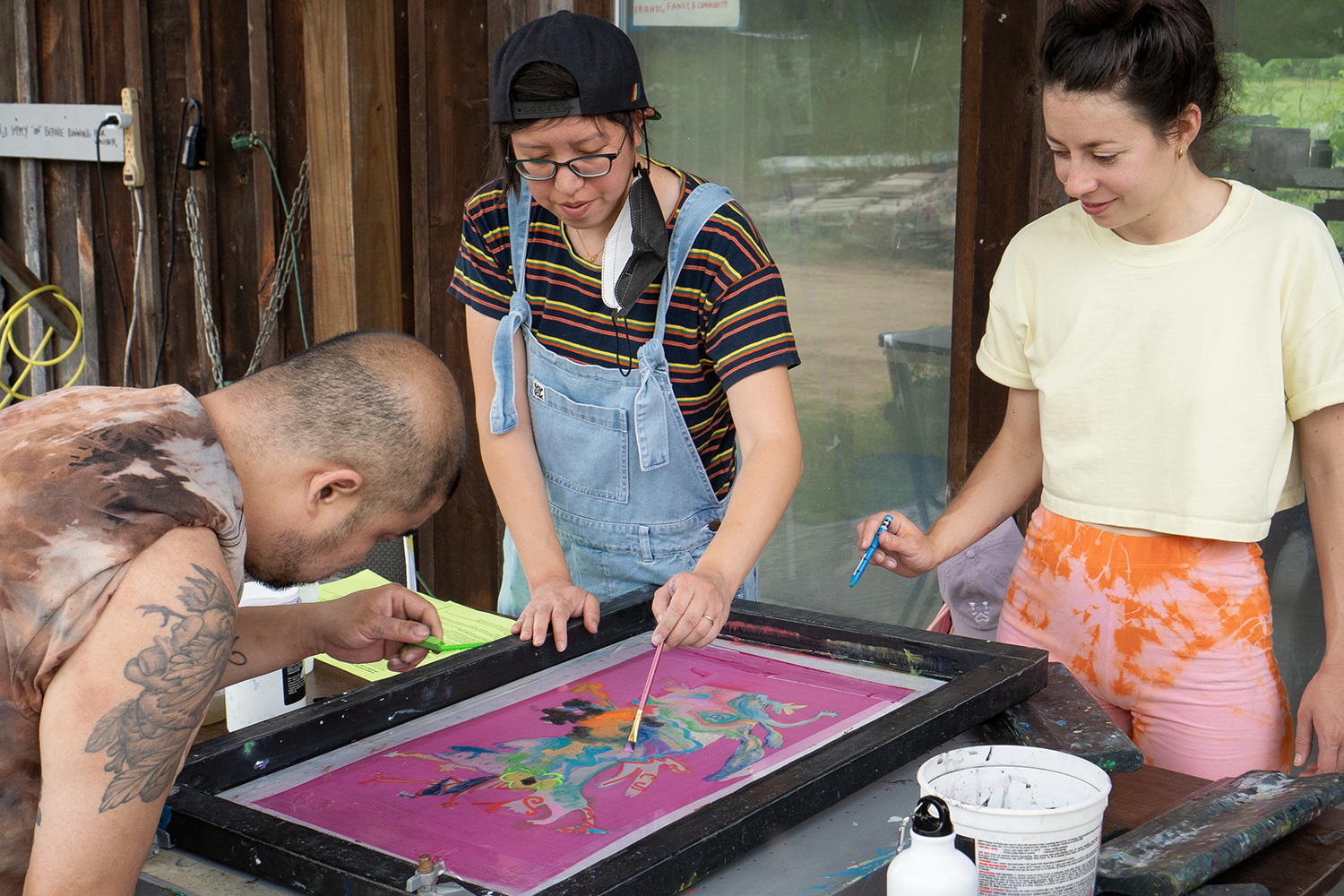 Three people stand around a table with a silkscreen printing frame. They are each adding paint to the screen.