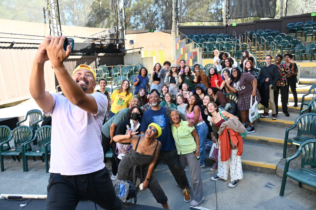 A person holds up a camera to take a selfie with a group of 20 people.