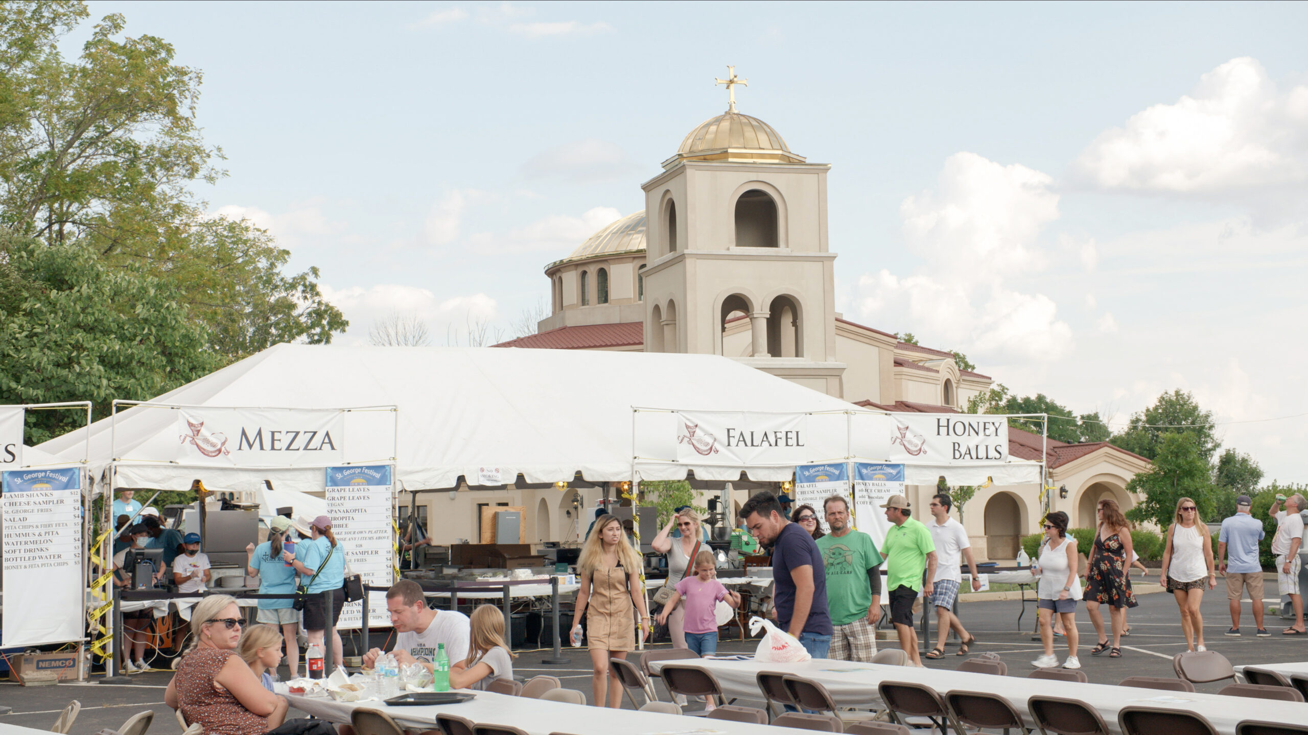 Festival goers stand in a line in front of a white tent and decorative Orthodox Church to get food like mezza, falafel, and honey balls