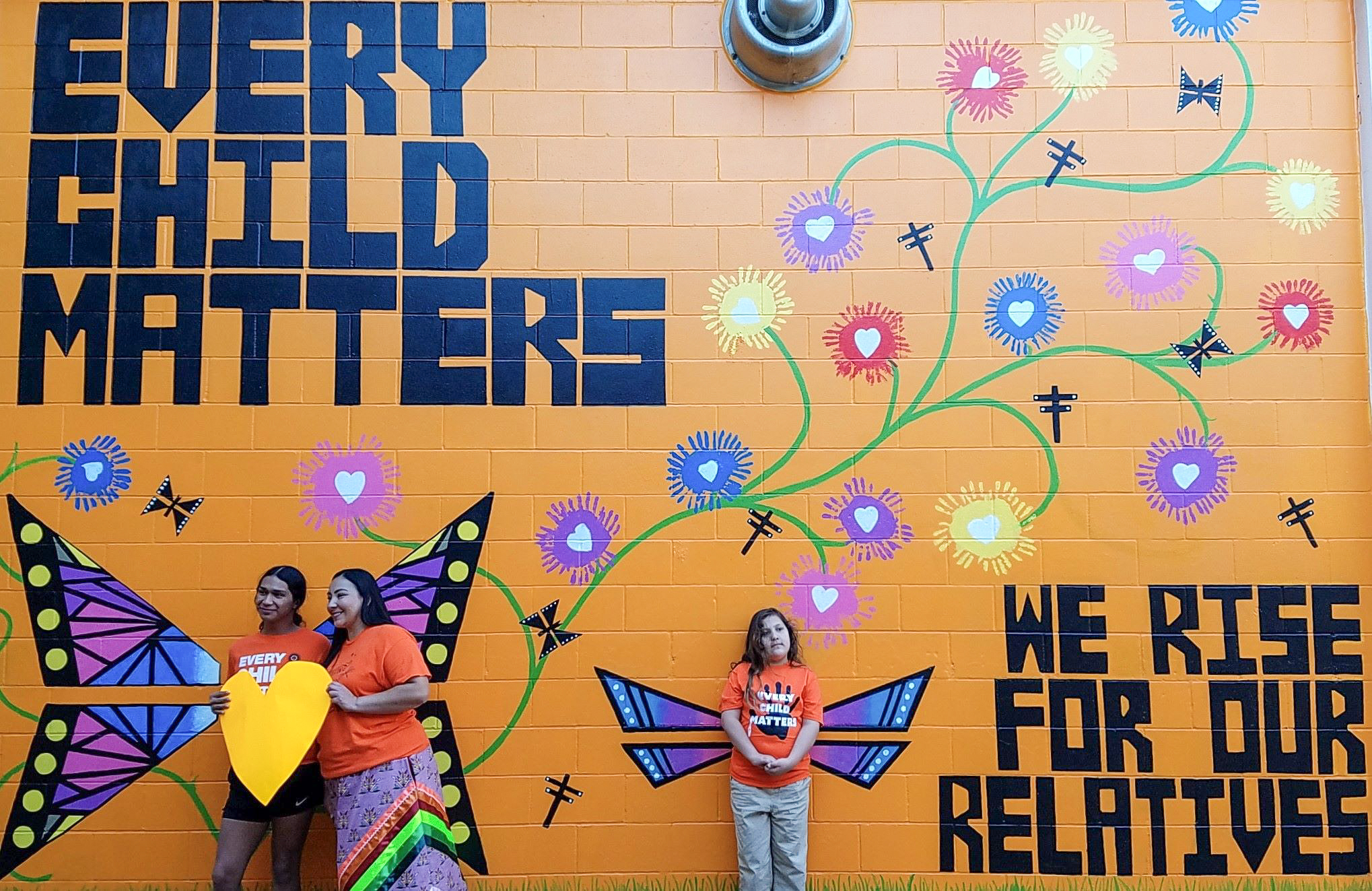 Two adults and one child stand by a large wall mural with a bright orange backdrop. The mural depicts flowers on vines extending towards the top, and words painted in black lettering that read, "every child matter" and "we rise for our relatives."