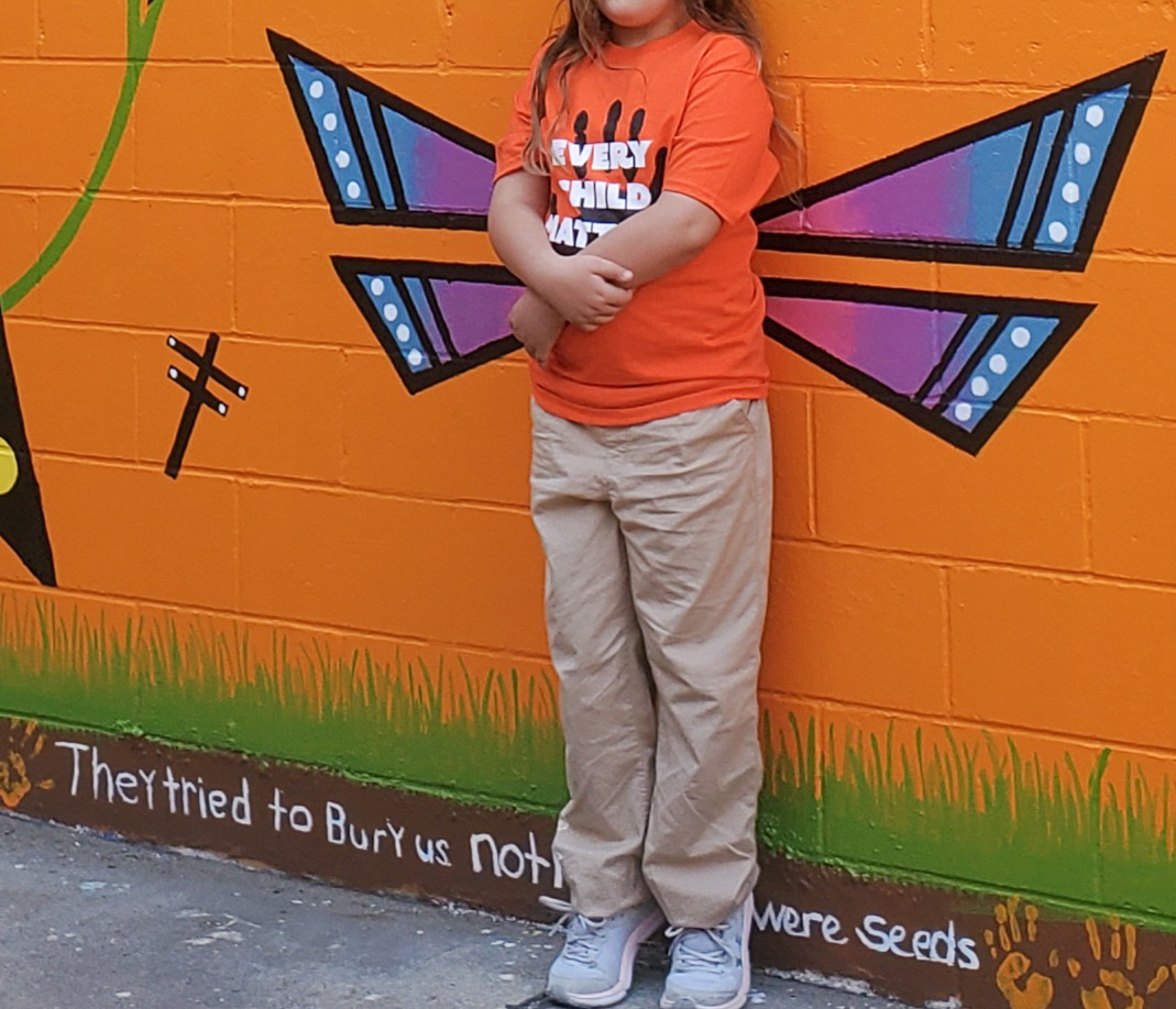 A child stands by a mural wall depicting dragonfly wings and words that read "They tried to bury us not knowing we were seeds."