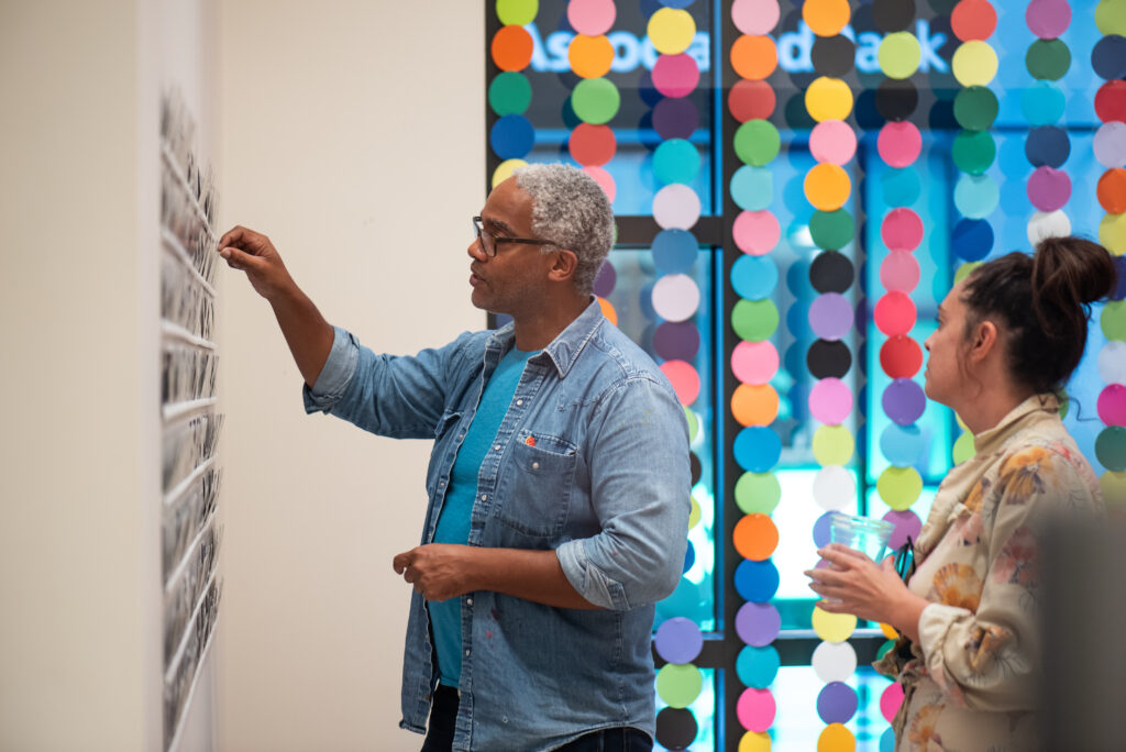 Two people look at a display on the wall, behind them is a colorful art instillation.