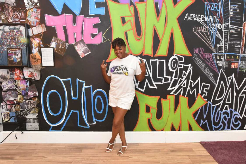 A person of dark skin tone wearing a white tee and shorts poses in front of a wall with different lettering and images. Some of the noticeable words in the background read "The Ohio Funk"