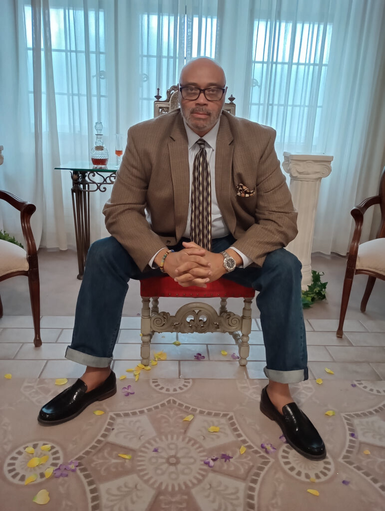 A person of light brown skin tone sits on a chair and poses slightly hunched over with their hands clasped in front of them. They are wearing large dark-rimmed glasses, a brown sport jacket, a pattern neck tie over a light shirt, blue denim jeans, and black loafers without socks.