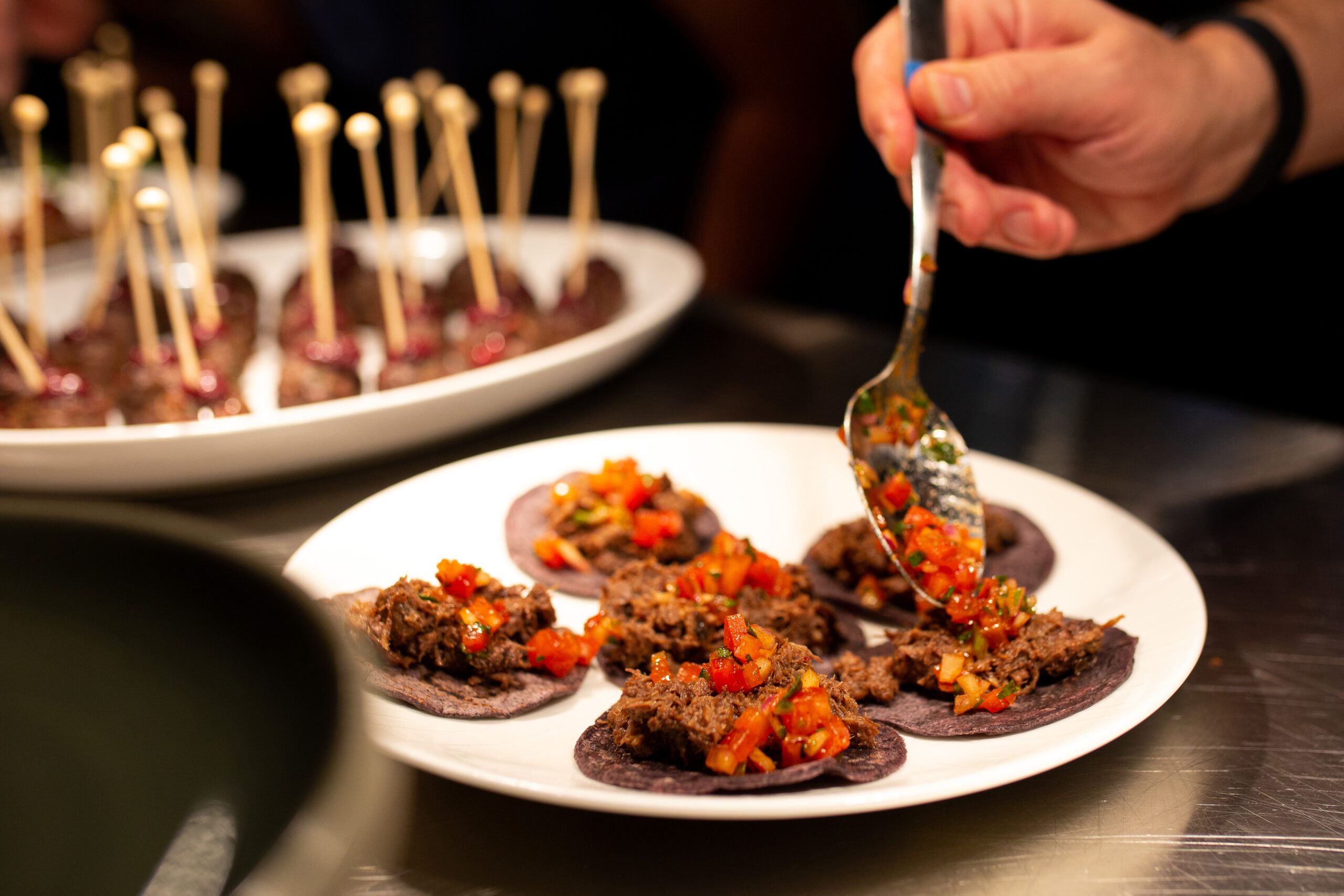 A chef uses a spoon to spread a garnish on a plate of bite size appetizers.